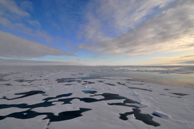 Parallel cloud bands converge at the horizon over a landscape of ice floes, melt pools, and a polynya to the right