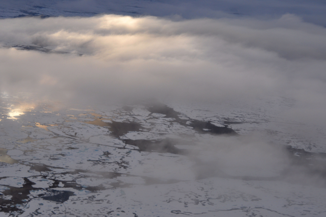 A helicopter eye's view of melting ice with clouds overhead illuminated througha layer of higher yet clouds