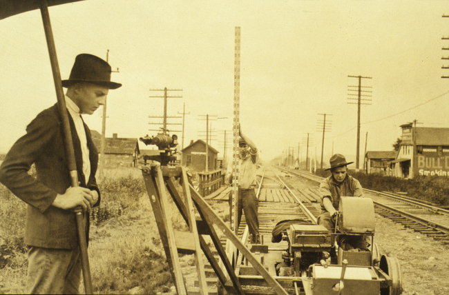 Recorder sitting while working with tabulating machine; umbrella man shadingleveling instrument; and rodman holding rod vertical over turning point