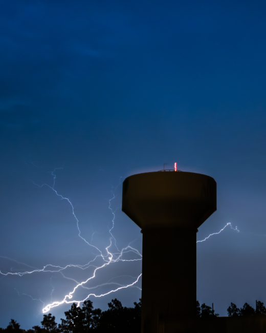 A strong lightning bolt coming from behind the trees; appearing to be reachingout for the water tower