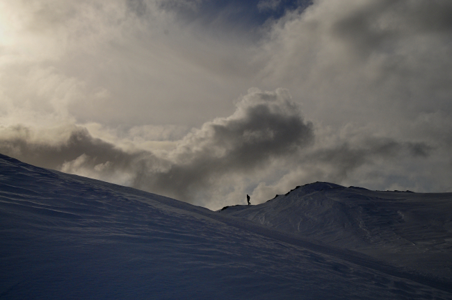 An AMLR scientist walks along the crest of a snow-covered mountainsilhouetted bya dramatic vista of clouds
