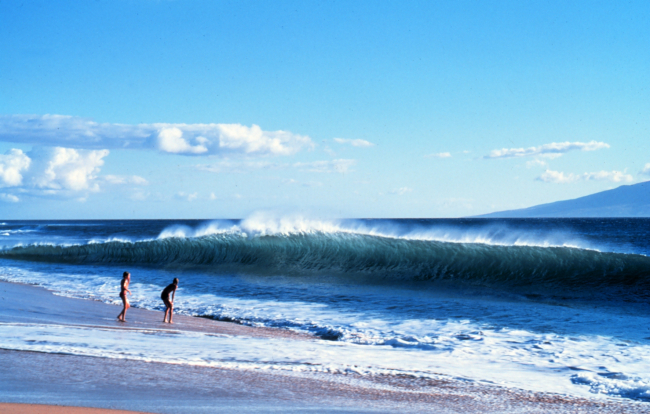 Two vacationers dwarfed by the shorebreak as an offshore wind whips spray offthe crest of the wave