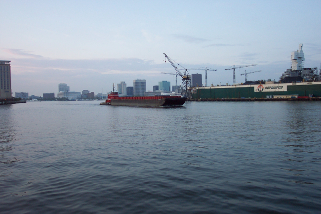 Tug pushing barge down the Elizabeth River with Norfolk skyline andshipyard in picture
