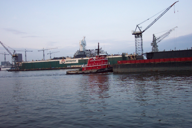 Tug pushing barge down the Elizabeth River with Navy ship in drydock