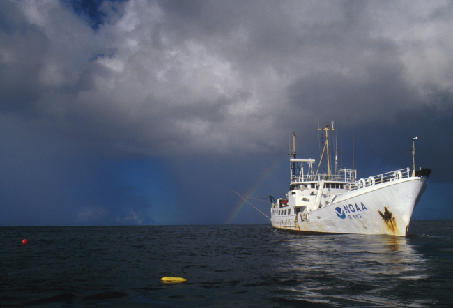 NOAA Ship TOWNSEND CROMWELL is rimmed by a rainbow whileit stands by during a deep diving operationwith down line and diver's PAM nearby