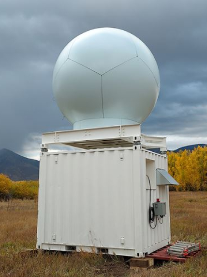 The X-Band, which has a globe-like shape on top and a square box shape on the bottom, sits outside in an open grassy area with views of the trees and mountains in the back. 