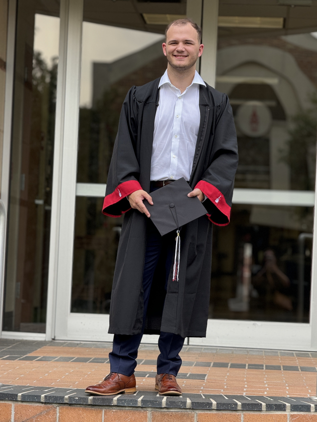 Christian posing in graduation robes and holding a graduation cap.