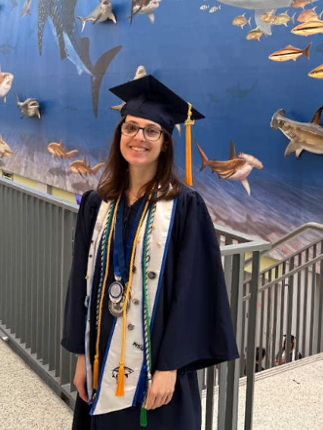 Courtney posing in a graduation cap and gown in front of an ocean mural.