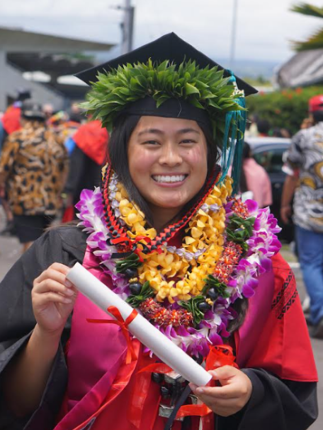 Michaela holding a rolled up degree in her graduation cap and gown. She is also wearing several flower leis around her neck and a leaf lei around her cap.