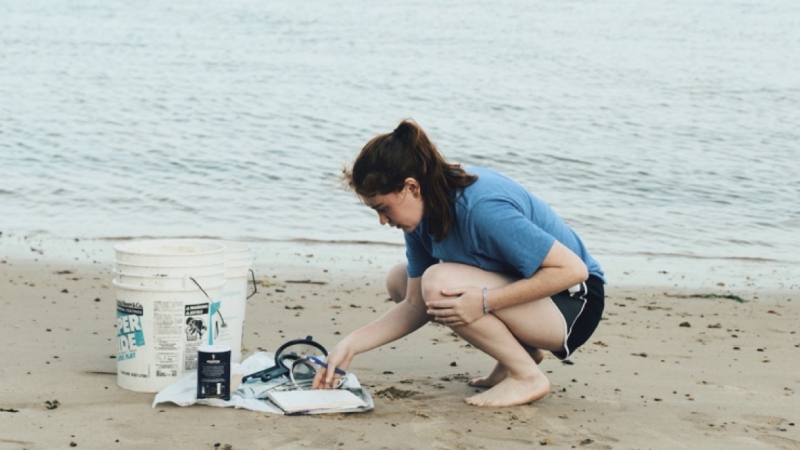 Leah crouches on a beach, organizing scientific sampling equipment.