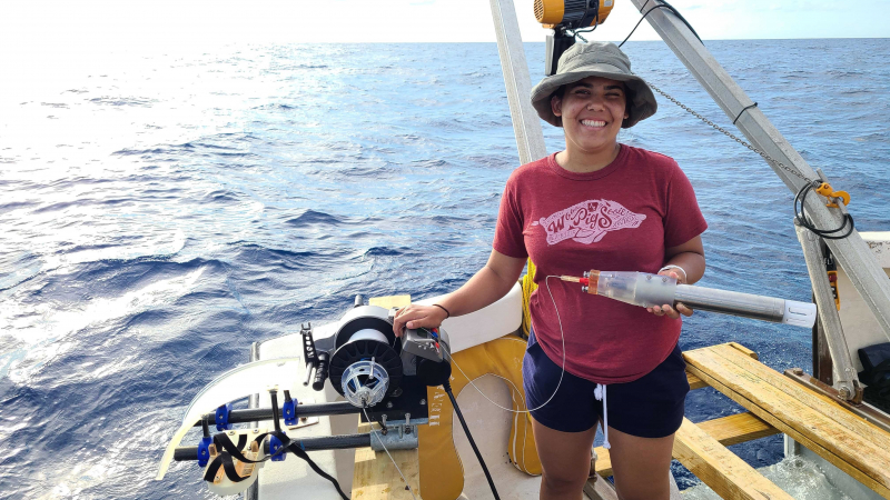 Adriana stands on a boat, holding a winch that she designed during the internship.