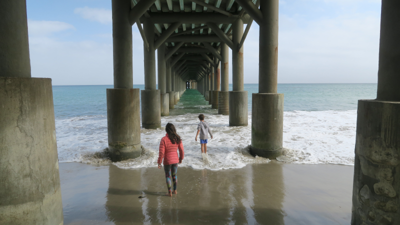 A young girl and boy run among the waves under a pier on a sandy beach. The perspective of the photo, taken from under the pier, shows a tunnel-like effect, where the two rows of columns holding up the pier create a tunnel. The children are facing away from the viewer, walking towards the waves between the columns.