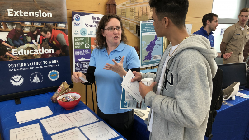 Grace Simpkins converses with a career fair attendee at a tabletop career event. A banner in the background has the words “extension” and “education” on it as well as the logos for NOAA, the Woods Hole Oceanographic Institution, and the Cape Cod Cooperative Extension. In the foreground is a table with sheets of paper and a bowl of candy.