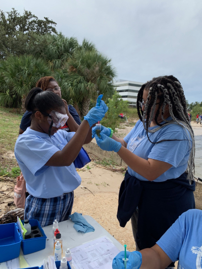 Students with protective goggles and gloves conduct water quality tests at Pontchartrain Beach with equipment and paperwork on the nearby table.