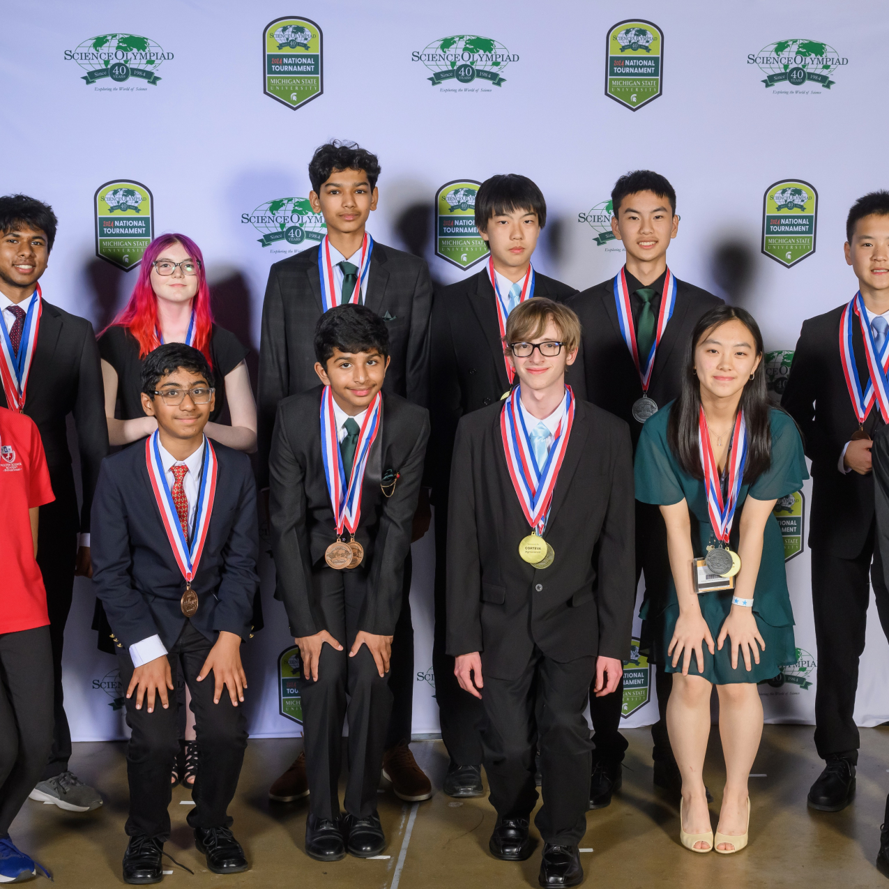 Twelve students dressed in formal attire pose for a group photo in front of a backdrop that has the 40th Science Olympiad National Tournament logo on it. 