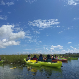 Students and educators in kayaks gather at the marsh edge.
