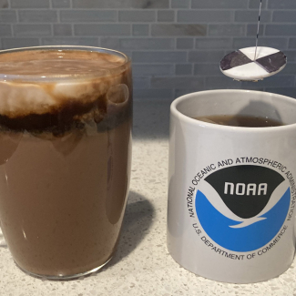 Two mugs are placed on a kitchen counter. One mug has the NOAA logo on it and a miniature secchi disk is suspended above it. (The secchi disk is made of a white disk with black on opposite quarters. It is hanging from a string that has markings every centimeter.) The other mug is made of clear glass, and it contains hot cocoa topped with a layer of espresso and a layer of milk. The top two layers are mixing, forming swirls.