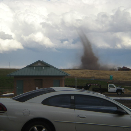 A slender tornado stretches from puffy white clouds to the ground. The funnel starts as light grey from the clouds and turns brown as it nears the ground, dirty with kicked up dirt and debris.