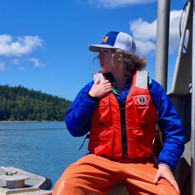 Devynn sits on a boat with her head turned to the side, looking off into the distance. She is wearing a life vest, rubber field pants, and other cold weather clothes.