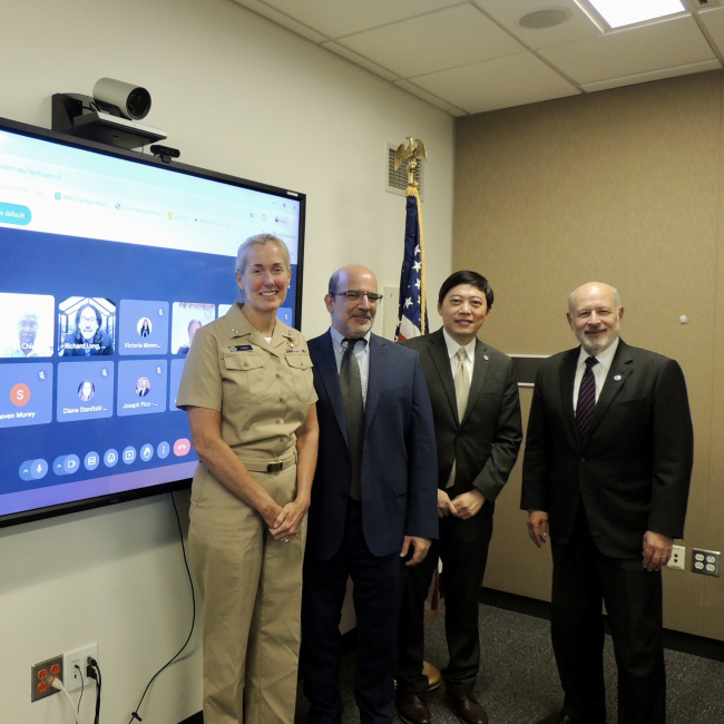 VADM Hann, Ben Friedman, Dr. Sen Chaio, and Dr. Rick Spinrad pose for a photo standing next to a virtual meeting that includes other members of NOAA and the EPP/MSI Cooperative Science Centers. The U.S. flag is behind them. 