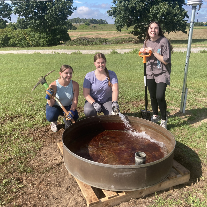 Three women and a large steel pool of water the size of a kiddie pool in front of them surrounded by a flat rural landscape.  Gabby is holding a pickaxe, the woman in the center holds a hose that is filling the pool, and the other holds a shovel. All three smile at the camera.