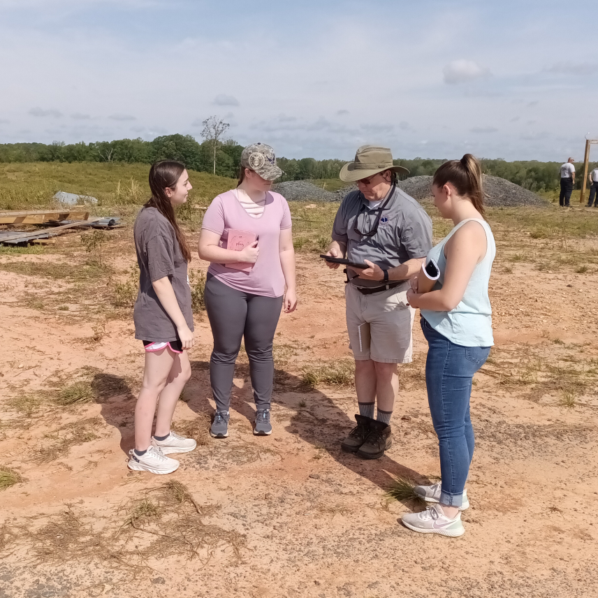 A group of four people stand in a flat rural area. A man at the center of the group wearing a National Weather Service shirt holds a tablet. Three women stand around and look at the tablet. 