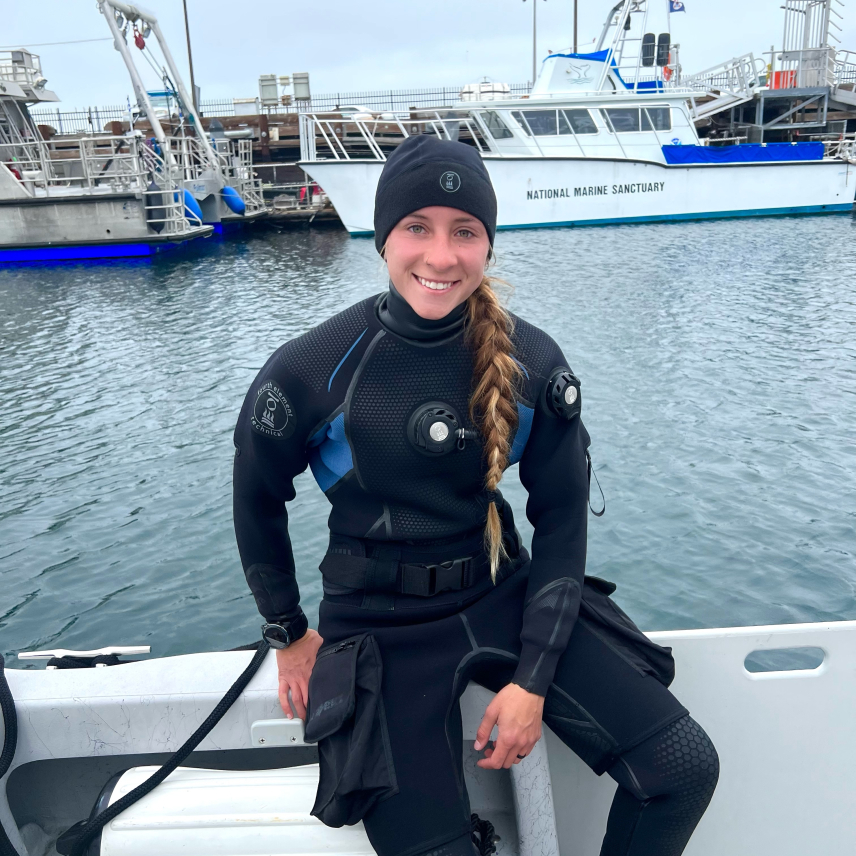 Gracia sits on the side of a small boat wearing a dive suit and smiling at the camera. Another boat in the background is labeled with text that reads "National Marine Sanctuary."