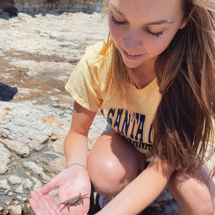 Caroline kneels on a rocky intertidal area and looks down at a small crab that rests in the palm of her hand.