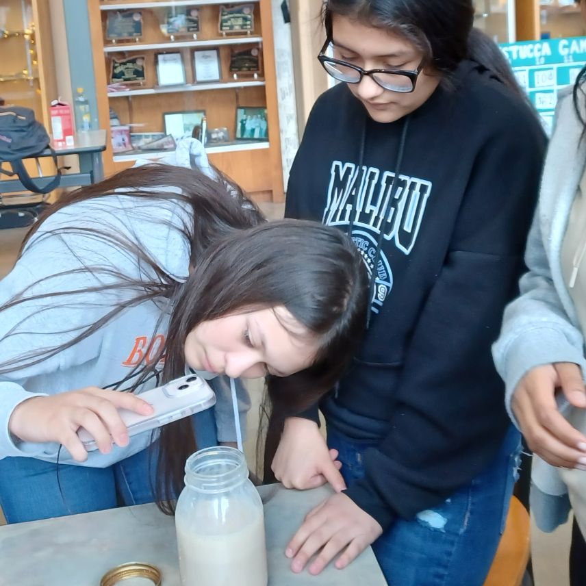 Two students stand over a mason jar of foamy milk. One student is bent over holding a cell phone to take pictures of the foam.
