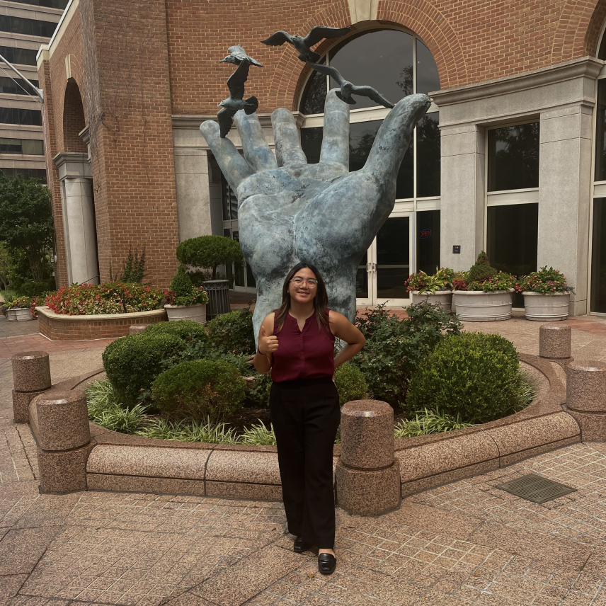 Roxanne gives a thumbs up in front of a statue of a large open hand that looks like it is releasing birds.