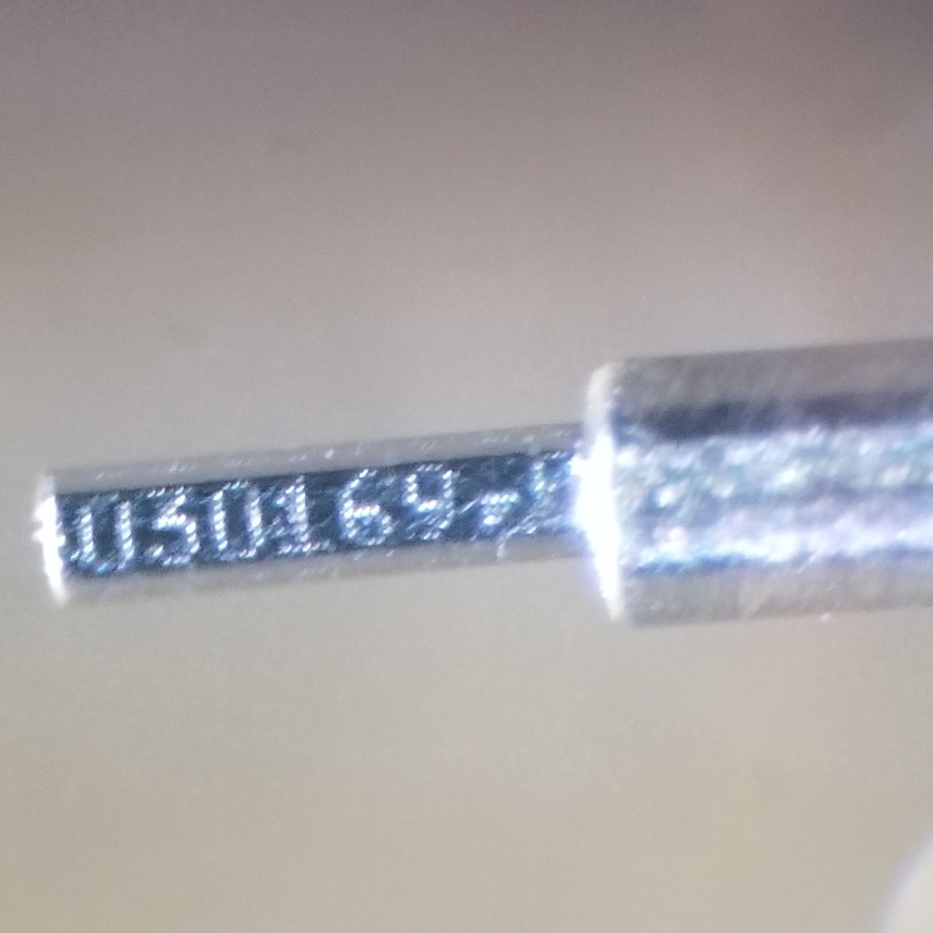 A view through a microscope of a thin metal bar with numbers etched into it. It is about half the width of the forceps that hold it.